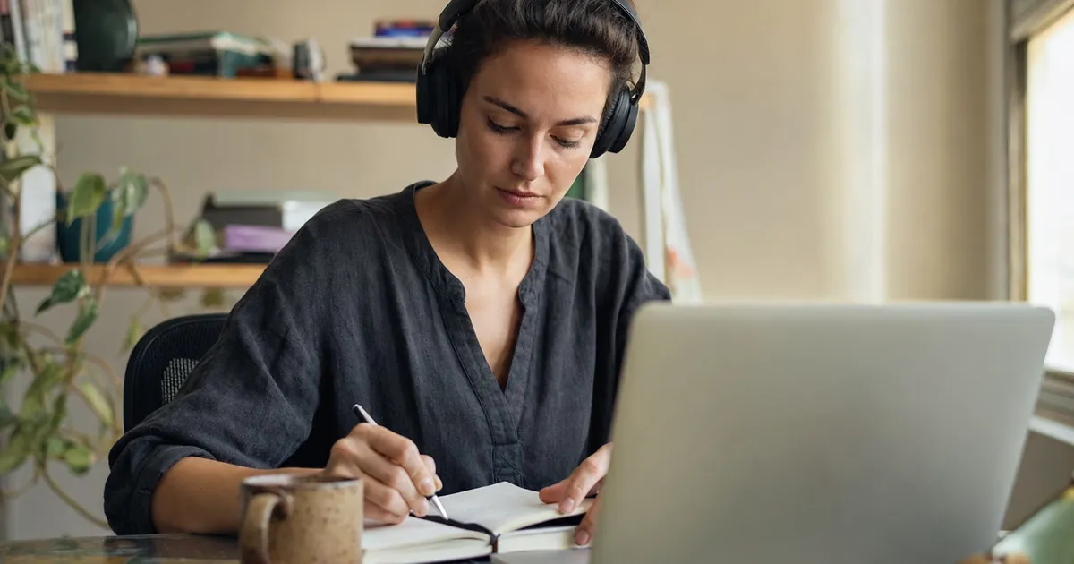 Kulaklıkla laptoptan online ders izleyip not alan bir kadın