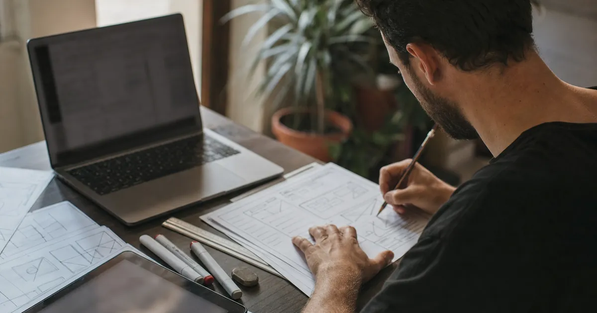 Kâğıt üzerine elle web sitesi taslağı çizen bir tasarımcı, yanında laptop