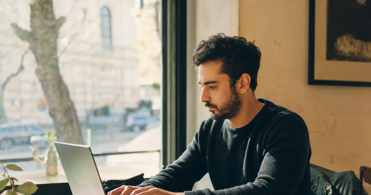 Kafe penceresinin önünde laptopla çalışan, yanında not defteri olan bir adam