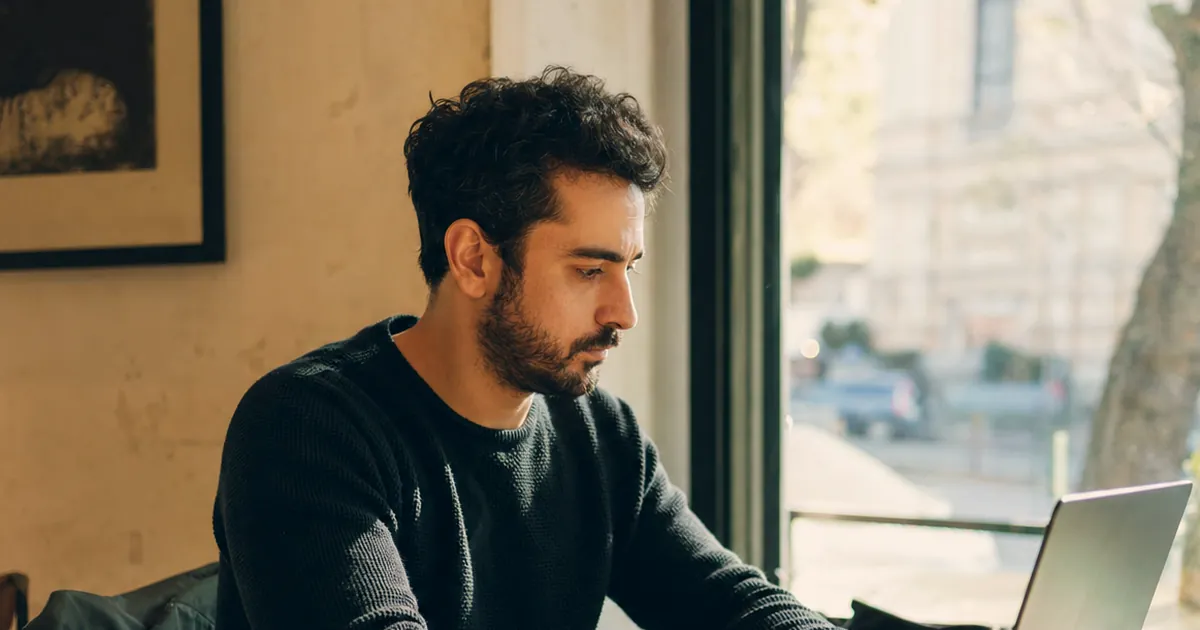 Kafe penceresinin önünde laptopla çalışan, yanında not defteri olan bir adam